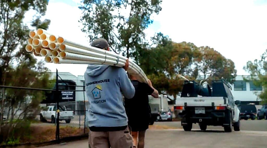 Two people carry a bundle of long metal pipes along a street, with a pickup truck in the background.